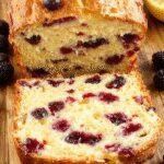 A freshly sliced Lemon Blueberry Yogurt Loaf on a wooden cutting board, garnished with fresh blueberries and lemon zest, with a soft crumb visible inside.